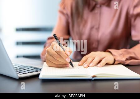 Mains d'une femme écrivant des notes dans un cahier en papier ouvert avec un stylo, assise à un bureau à côté d'un ordinateur portable, représentant la planification, l'organisation et la productivité professionnelle dans un environnement de bureau Banque D'Images
