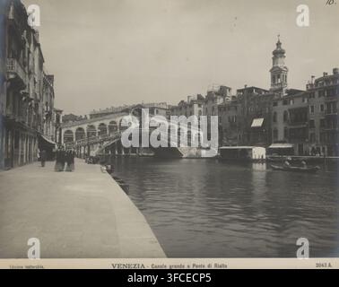 Venezia - Canale grande e Ponte di Rialto, Vue du Pont du Rialto à Venise, photographie, numéro : '3043 A.', numéro : '10.', photographe : Neue Photographische Gesellschaft, Venise, c. 1900, papier, carton, tirage argentique au gélatine, tirage argentique au gélatine développé, hauteur 194 mm x largeur 243 mm, canal, pont, gondole, Pont du Rialto., Une scène riveraine en niveaux de gris montre une promenade en pierre à côté d'un large canal avec une eau calme et ondulante, un grand pont en arc traversant le canal avec des balustrades ornées et plusieurs arches, des rangées de bâtiments à plusieurs étages avec balcons et façades variées bordant les deux côtés Banque D'Images