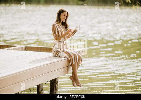 Une femme savoure un moment de détente, lisant au bord du lac paisible tout en sirotant une boisson chaude. La lumière du soleil danse sur l'eau. Banque D'Images