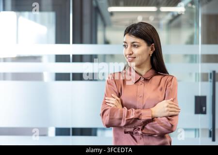 Jeune femme d'affaires professionnelle debout les bras croisés, regardant au loin et souriant dans un bureau d'entreprise moderne, représentant la confiance, le succès et le leadership Banque D'Images