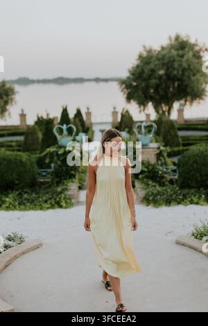 Une jeune femme dans une robe jaune fluide marche avec bonheur à travers un jardin luxuriant au coucher du soleil. Banque D'Images