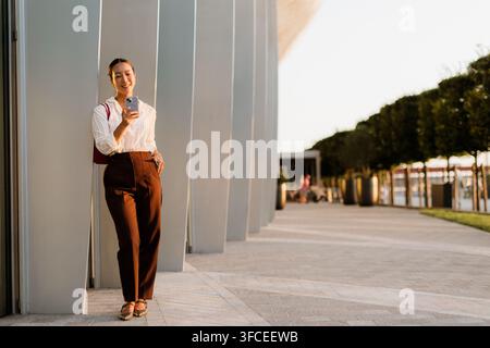 Une jeune femme coréenne se tient avec assurance sur une passerelle moderne, souriant et utilisant son smartphone. La lumière chaude du coucher de soleil met en valeur sa tenue élégante. Banque D'Images