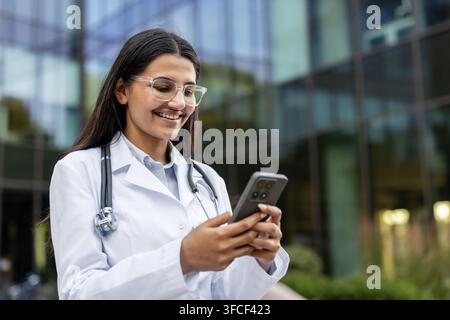 Médecine indienne portant une blouse de laboratoire et un stéthoscope. Souriante en utilisant un smartphone pour une consultation à distance ou en communiquant via une application de santé devant un bâtiment médical moderne Banque D'Images