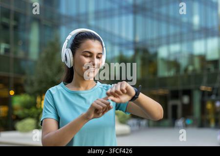 Jeune femme athlétique souriant et vérifiant sa montre intelligente, portant des écouteurs tout en se tenant en plein air dans un environnement urbain, suivant ses progrès de fitness et profitant de la musique pendant son entraînement Banque D'Images
