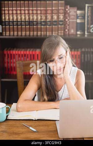 Jeune femme fatiguée avec des lunettes repose sa joue sur sa main à côté d'un ordinateur portable, d'un agenda ouvert, d'un stylo et d'une tasse de café tout en étudiant à la maison. Banque D'Images