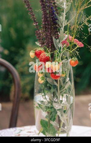 Des fraises fraîches et des herbes fleuries forment un arrangement botanique rustique dans un vase en verre transparent, créant une nature morte inspirée du jardin. Banque D'Images