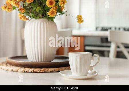 Tasse à café blanche à côté d’un vase avec des fleurs orange dans une cuisine lumineuse, intérieur automnal et confortable pour le café du matin, décoration saisonnière et vie lente. Banque D'Images