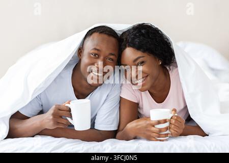 Heureux couple noir couché sous couverture, buvant du thé chaud, se réchauffant par temps froid. Amoureux afro-américains joyeux avec des tasses restant dans le lit d Banque D'Images