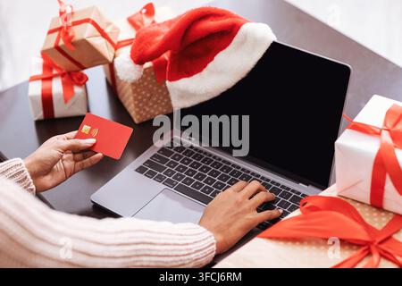 Préparation pour Noël. Vue par-dessus l'épaule d'une femme afro-américaine tenant une carte de crédit et utilisant un ordinateur avec un écran noir vide pour un modèle Banque D'Images