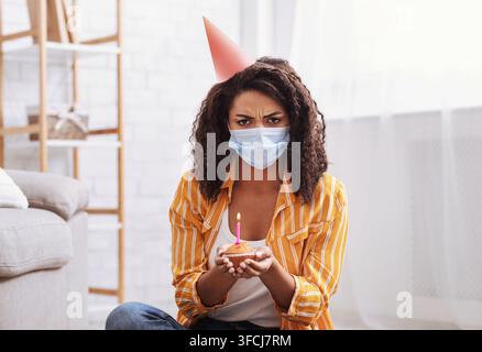 Femme noire triste portant un chapeau de fête et un masque médical, célébrant son anniversaire seule en quarantaine ou en auto-isolement. Femme afro-américaine en colère regardant la caméra Banque D'Images