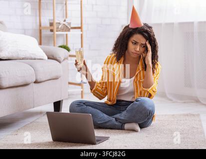 Femme noire triste et contrariée assise par terre, tenant un verre de vin, portant un chapeau de fête. Dame noire célébrant son anniversaire seule et en ligne. Concept de solitude Banque D'Images