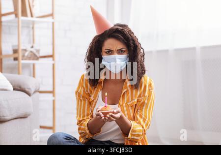 Quarantaine, auto-isolement et pandémie. Femme afro-américaine en colère et frustrée regardant la caméra, tenant un gâteau avec une bougie, portant un chapeau de fête et Pr Banque D'Images