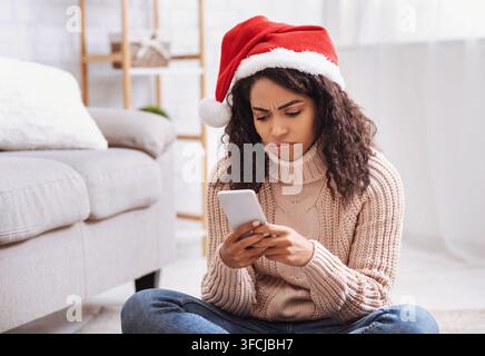 Femme afro-américaine frustrée utilisant un téléphone portable assise par terre dans le salon. Dame noire confuse et en colère célébrant seule, attendant son ca Banque D'Images