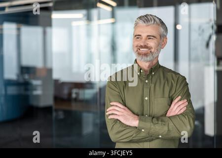 Homme d'affaires mûr et confiant aux cheveux gris et à la barbe, portant une chemise verte, souriant et regardant au loin tout en se tenant les bras croisés dans un bâtiment de bureaux moderne Banque D'Images