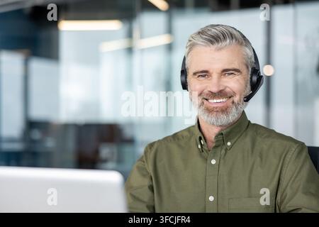 Homme barbu mûr souriant avec bonheur et portant un casque avec microphone, fournissant un support technique ou un service client en ligne dans un environnement de bureau moderne Banque D'Images