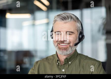 Homme mûr à la barbe grise souriant dans un casque et un microphone, fournissant un support en ligne amical et un service client virtuel depuis un environnement de bureau moderne, agent confiant et professionnel Banque D'Images