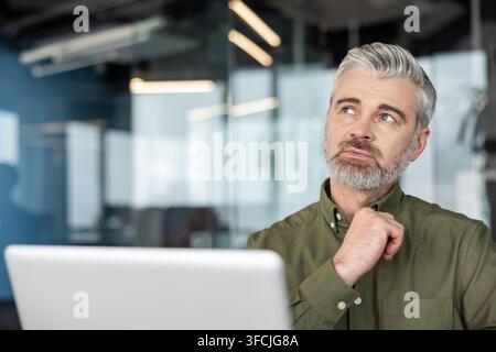 Homme d'affaires mûr aux cheveux gris dans un bureau, assis devant un ordinateur portable, regardant vers le haut dans une réflexion profonde tout en planifiant, brainstormant et résolvant des problèmes avec une confiance concentrée Banque D'Images