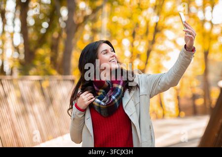 Belle femme millénaire dans une tenue tendance avec selfie sur mobile téléphone au parc jaune d'automne Banque D'Images