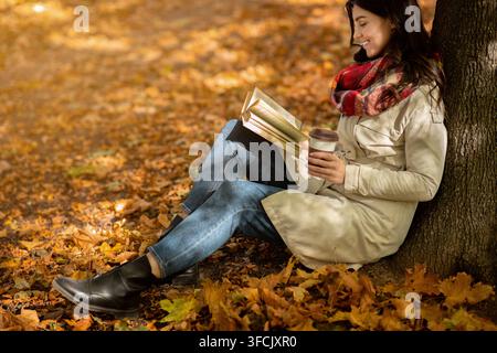 Belle jeune femme dans un manteau élégant se détendant sur des feuilles d'automne avec un livre captivant au parc, espace de copie Banque D'Images