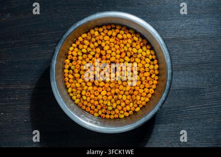 Vue de dessus de baies de l'épine-vierge orange mûres flottant dans l'eau dans un bol en métal sur un fond en bois sombre, gros plan. Concept de lavage de l'automne frais Banque D'Images