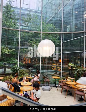 Des personnes assises à des tables avec des ordinateurs portables et des livres dans un café moderne en atrium vitré doté d'une grande lampe de sol sphérique et d'un jardin intérieur Banque D'Images