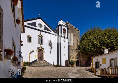 Église et murs du château médiéval à Óbidos, Portugal. Une église blanchie à la chaux et des fortifications en pierre se dressent le long d'une ruelle tranquille. Banque D'Images