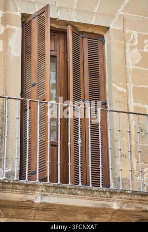 Volets en bois ouverts et un balcon en fer forgé encastré dans une façade en pierre patinée, montrant des détails architecturaux traditionnels à Baeza, en Espagne. Banque D'Images