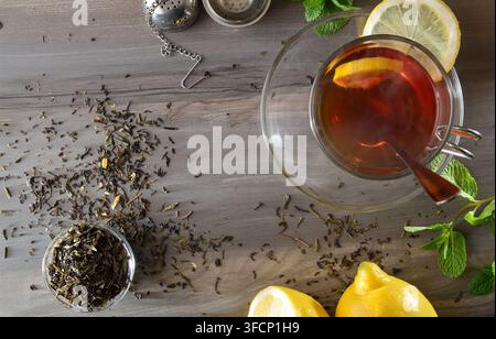 Une tasse à thé transparente repose sur une soucoupe en verre sur une table en bois sombre avec des ingrédients tels que des citrons et de la menthe fraîche, ainsi qu'un bol de feuilles de thé séchées. En haut Banque D'Images