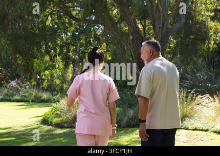Aidante féminine marchant avec un homme âgé en polo beige dans le jardin, portant une tenue d'infirmière rose et des montres Banque D'Images