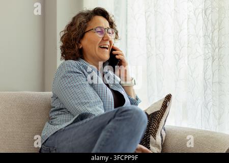 Femme mûre heureuse riant pendant un appel téléphonique sur le canapé Banque D'Images