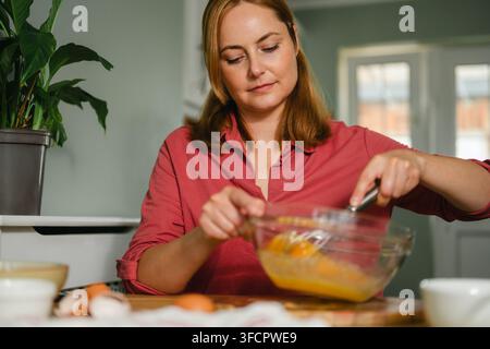Femme dans la cuisine fouettant des œufs frais dans un bol en verre, pâtisserie maison Banque D'Images