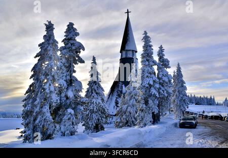 Une église en bois traditionnelle avec un clocher élevé se dresse entourée de pins lourdement chargés de neige sous un ciel d'hiver dramatique. Des voitures garées le long d'une Banque D'Images