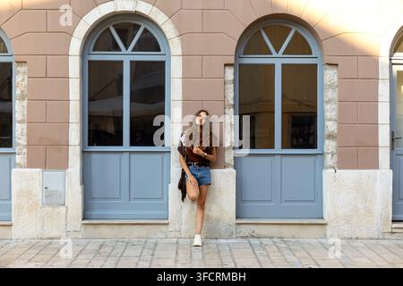 Jeune femme posant entre des fenêtres en arc sur une façade pêche. Jeune femme aux cheveux longs et bouclés posant entre des fenêtres en arc sur la façade d'un bâtiment pêche. Banque D'Images