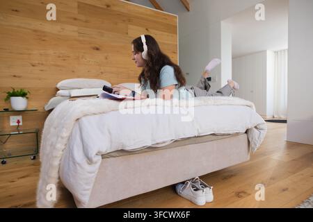 Femme allongée sur le ventre sur un lit dans une chambre, portant des écouteurs et tenant un appareil Banque D'Images