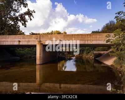Pont en béton sur une rivière calme traversant un paysage verdoyant avec des nuages blancs duveteux dans le ciel bleu Banque D'Images