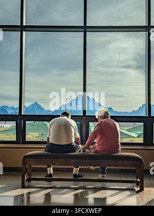 Deux visiteurs sont assis sur un banc à l'intérieur du Jackson Lake Lodge, encadrés par des fenêtres du sol au plafond et les sommets lointains de la chaîne des Grand Teton. Banque D'Images