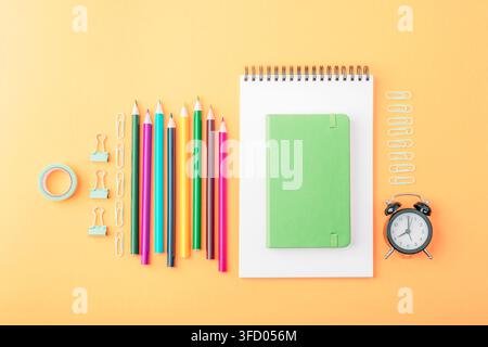 Composition statique avec des crayons colorés, un cahier vierge, une règle, des trombones et un réveil sur fond orange. Concept retour à l'école. Vue supérieure Banque D'Images