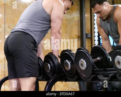 Profil latéral concentré d'un homme athlétique s'appuyant sur un rack à haltères lourd tout en regardant son reflet dans le miroir de la salle de sport. Illustre la concentration, l'esprit-Mu Banque D'Images