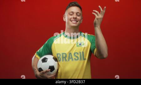 Homme tient le ballon de football sous le bras et retourne les doigts en studio tout en portant le maillot brésilien et en souriant avec les yeux fermés ; célébration de fierté. Banque D'Images
