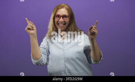 Sénior femme pointe ses deux index vers le haut et sourit largement, portant des lunettes rouges et une chemise à carreaux clairs en studio ; célébration de joie. Banque D'Images