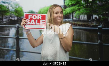 La femme pointe le doigt à pour le signe de vente sur la rampe de canal de rue tout en tenant l'écriteau blanc et souriant ; promotion de la confiance des ventes. Banque D'Images