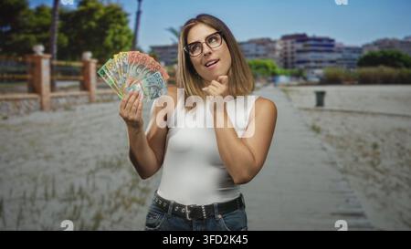 Femme tenant un éventail de billets de dollars australiens et touchant son menton dans la rue ; curiosité pensive sur l'argent. Banque D'Images