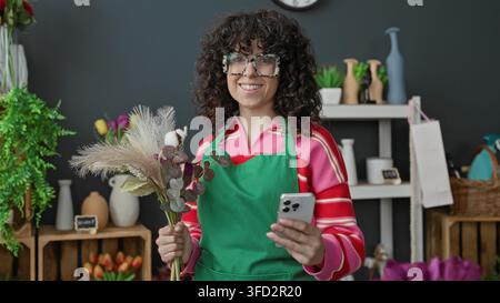 Femme fleuriste dans un tablier vert tenant un bouquet séché et regardant son smartphone au milieu de vases et d'étagères dans un studio de magasin de fleurs ; busi local joyeux Banque D'Images