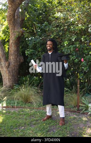 Homme africain tenant un parchemin de diplôme et une toge, vêtu d'une robe noire, souriant dans un jardin Banque D'Images