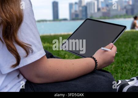 Jeune femme tenant une tablette avec un écran sombre vierge et un stylet tout en étant assise en plein air près du bord du lac de Chicago, avec l'horizon urbain légèrement flou Banque D'Images