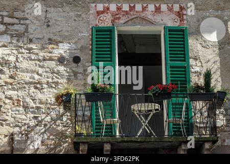 un balcon européen traditionnel sur une vieille maison en pierre. Le balcon présente des volets en bois verts et vibrants ouverts, une table en fer forgé et deux chaises. Banque D'Images
