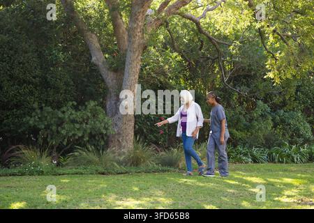 Sénior femme en sandales, aide-soignante en blouse grise marchant sur la pelouse en touchant un arbuste rose, espace libre Banque D'Images