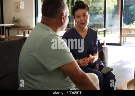 Infirmière asiatique en blouse bleu marine tenant une bouteille de pilules et une tablette, parlant à un client sur un canapé, espace libre Banque D'Images