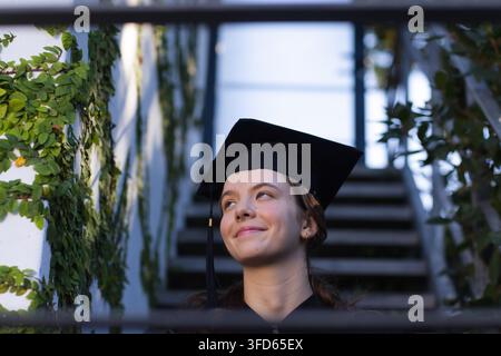 Femme adulte debout sur des escaliers extérieurs portant une coiffe et une robe de graduation, encadrée par des barreaux et des vignes, souriante Banque D'Images