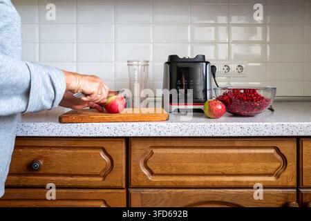 Une femme coupe une pomme sur une planche en bois dans une cuisine avec un mixeur et un bol de canneberges sur le plan de travail. Banque D'Images